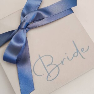 May include: A white gift box with a blue satin ribbon tied in a bow. The box has the word "Bride" written on it in a light blue cursive font.