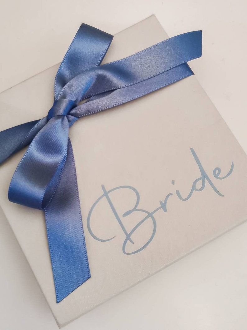May include: A white gift box with a blue satin ribbon tied in a bow. The box has the word "Bride" written on it in a light blue cursive font.