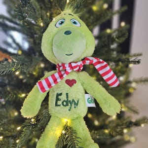 May include: A plush green character doll with a red and white striped scarf. The doll has a red heart and the name "Edyn" embroidered on its belly. The doll is set against a backdrop of a Christmas tree.