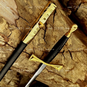 May include: A decorative sword with a gold-colored hilt and crossguard, and a black handle. The sword's blade is silver. The sword is placed on a piece of wood. The sword's sheath is black with gold accents.