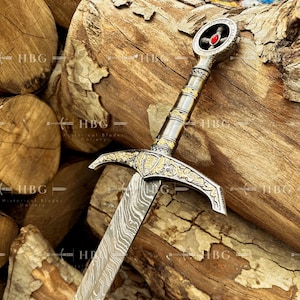 May include: A detailed image of a decorative sword with a silver and gold handle, a cross-shaped pommel with a red gem, and a patterned blade. The sword rests against a backdrop of wooden logs.