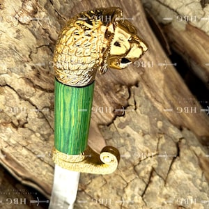 May include: A decorative dagger with a gold lion head handle and a green and gold wooden grip. The blade is silver. The dagger is displayed on a wooden surface.