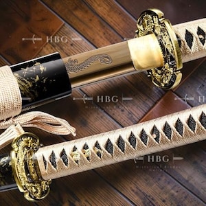 Könnte beinhalten: Nahaufnahme eines japanischen Katana-Schwertes. Der Griff ist goldfarben und der Griff beige. Die Klinge hat eine Drachengravur. Die Scheide ist schwarz mit goldenen Details. Das Schwert liegt auf einer Holzoberfläche.