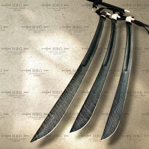 K&ouml;nnte beinhalten: Drei schwarze Schwerter mit komplizierten Mustern auf den Klingen. Die Schwerter sind f&auml;cherf&ouml;rmig auf einem hellbraunen Hintergrund angeordnet.