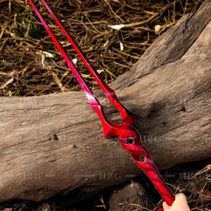 May include: A red spear with a unique design, featuring two long, parallel blades that converge at the base. The spear has a glossy finish and is held by a hand, with a wooden log in the background.
