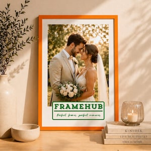 May include: An orange framed print showing a couple on their wedding day. The print has the text "FRAMEHUB" and "Perfect frames. perfect memories" in a green rectangle. The print is displayed on a wooden surface with decorative items, including books and a candle.