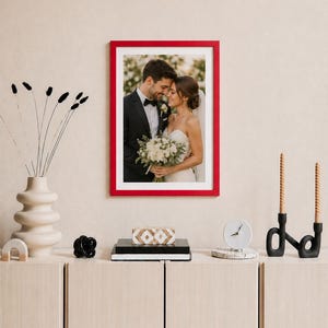 May include: A framed wedding photograph with a red border is displayed on a wall. The image shows a couple embracing, the bride holding a bouquet. Below, a white vase, decorative objects, and a clock sit on a light-coloured cabinet.
