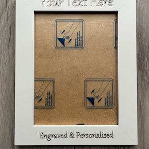 Op de afbeelding: Een witte fotolijst met de tekst "Your Text Here" bovenaan en "Engraved & Personalised" onderaan. De lijst heeft een rechthoekige opening en bevat instructies om de beschermfolie te verwijderen.