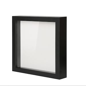 May include: A black shadow box frame with a clear glass front and a white backing. The frame is square and has a thick, solid border. The shadow box is designed to display items.