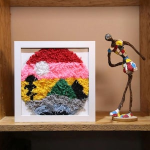 May include: A framed, circular textile artwork depicting a landscape. The piece uses red, pink, yellow, green, and grey colours. A small, abstract sculpture of a person is positioned to the right of the artwork.