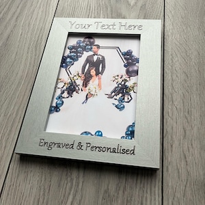 Op de afbeelding: Zilveren fotolijst met een trouwfoto. De lijst heeft de tekst "Your Text Here" bovenaan en "Engraved & Personalised" onderaan. De foto toont een bruidspaar met blauwe ballonnen.