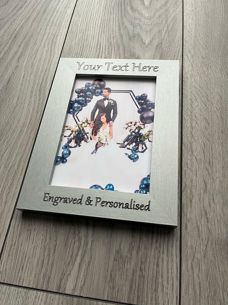 Op de afbeelding: Zilveren fotolijst met een trouwfoto. De lijst heeft de tekst "Your Text Here" bovenaan en "Engraved & Personalised" onderaan. De foto toont een bruidspaar met blauwe ballonnen.