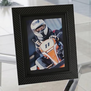 May include: A rectangular picture frame with a black carbon fiber texture finish containing a photograph of a person wearing a white helmet and racing gear while driving an orange go-kart with the number 11 displayed on the front panel.