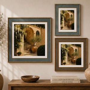 May include: Three framed wall art prints featuring a Mediterranean stone archway with cascading white flowers, a terracotta pot, and a staircase. The prints are displayed in blue and brown frames with white matting, arranged on a neutral wall.