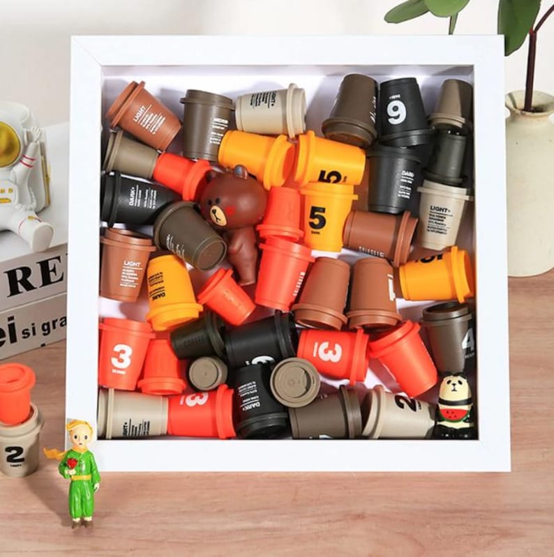 May include: A display of miniature coffee cup toys within a white framed shadow box. The cups are in shades of brown, orange, and black, with printed numbers and text. A small bear figurine is also included in the arrangement.