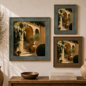 May include: Three framed wall art prints depicting a Mediterranean stone courtyard with arched doorways, stone stairs, and potted plants. The frames are blue and brown. Below, two stacked books titled Live Beautifully and This Is Home.