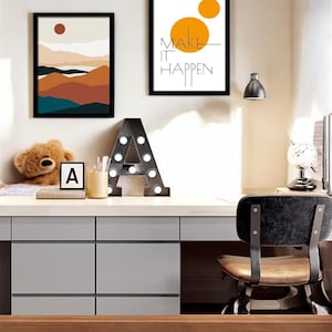 Könnte beinhalten: Zwei gerahmte Drucke an einer weißen Wand über einem Schreibtisch. Der Druck links ist eine Landschaft mit einer roten Sonne, braunen und blauen Bergen und einem weißen Himmel. Der Druck rechts ist weiß mit einem gelben Kreis und dem Text "MAKE IT HAPPEN".