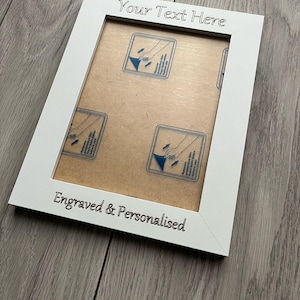 Op de afbeelding: Witte fotolijst met de tekst "Your Text Here" en "Engraved & Personalised" in zwart. De lijst heeft een lichtbruine achterkant met blauw-witte prints. De lijst staat schuin op een grijze houten ondergrond.