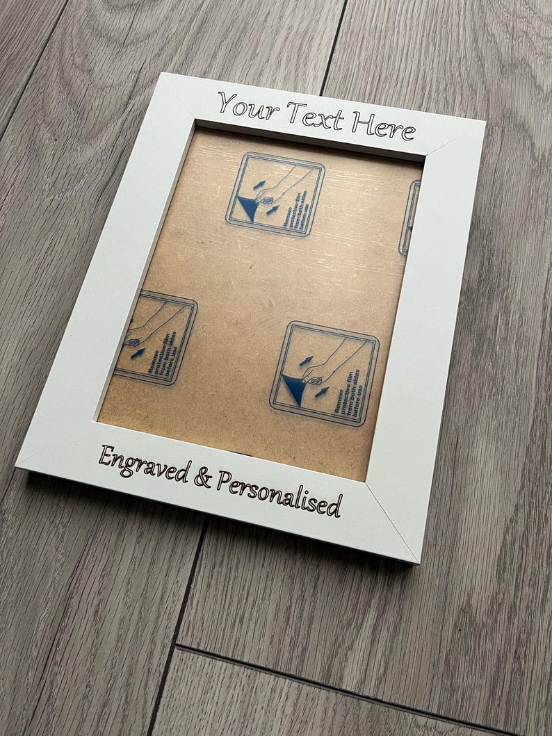 Op de afbeelding: Witte fotolijst met de tekst "Your Text Here" en "Engraved & Personalised" in zwart. De lijst heeft een lichtbruine achterkant met blauw-witte prints. De lijst staat schuin op een grijze houten ondergrond.