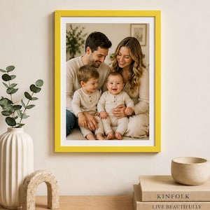 Puede incluir: Un retrato familiar enmarcado en amarillo. La fotografía muestra a una familia sonriente de cuatro: una madre, un padre y dos niños pequeños. La familia viste ropa de tonos neutros. El marco está colgado en una pared color crema.