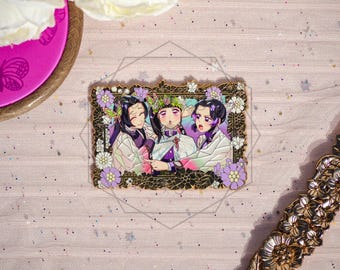 May include: A gold enamel pin depicting three anime-style characters within a decorative frame, adorned with purple and white floral details. The characters are clad in white and purple attire. The pin is set against a pink backdrop.