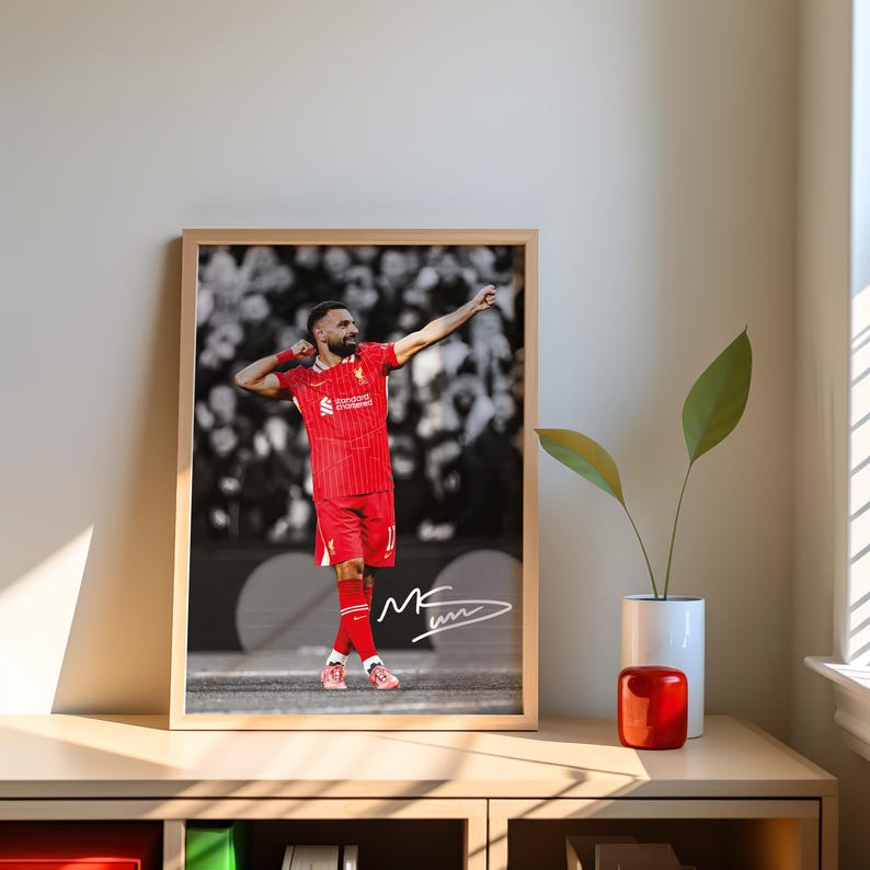 May include: Framed print of a footballer in a red kit, celebrating with arms raised. The background is a black and white crowd. The print includes a signature and is displayed on a wooden table. The frame is a light brown color.