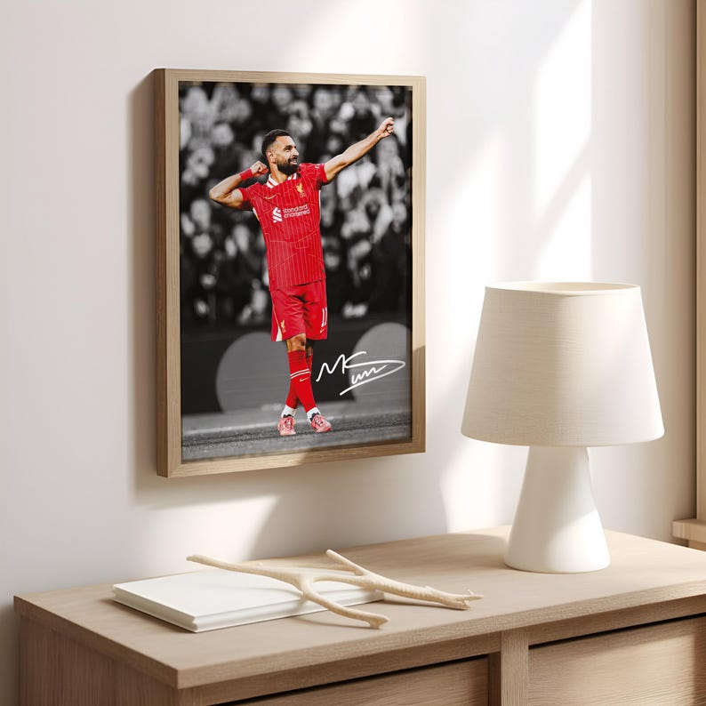 May include: Framed print of a soccer player in a red kit, celebrating with an arm raised. The background is a monochrome image of a crowd. The print is in a light brown wooden frame and includes a signature.