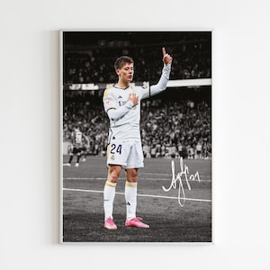 May include: A black and white photo of a soccer player wearing a white jersey with a gold trim and the number 24 on the front. The player is raising his right hand in the air and has a pink shoe on his right foot. The player is standing on a soccer field with a crowd of people in the background. The photo is signed with the name "Aylin".