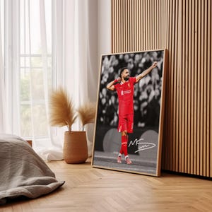May include: Framed print of a footballer in a red kit, celebrating a goal. The background is monochrome, contrasting with the player's red uniform. Includes a signature. The frame is light wood. The print is displayed indoors.