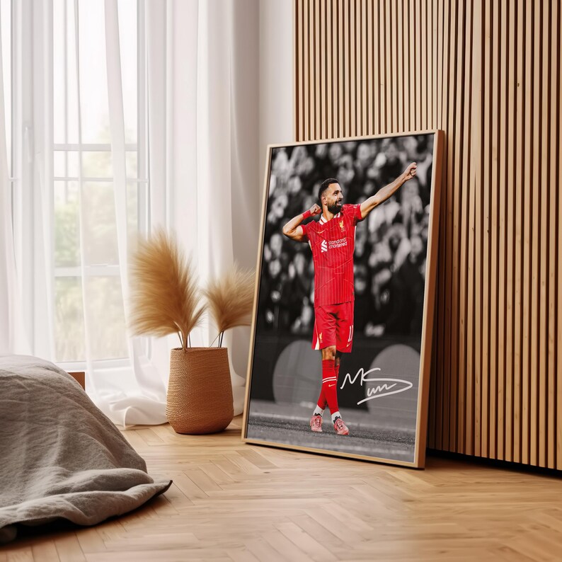 May include: Framed print of a footballer in a red kit, celebrating a goal. The background is monochrome, contrasting with the player's red uniform. Includes a signature. The frame is light wood. The print is displayed indoors.