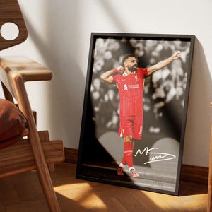 May include: Framed print of a footballer in a red kit, celebrating with arms raised. The background is a blurred black and white image. The print is in a black frame, with a signature in the bottom right corner.