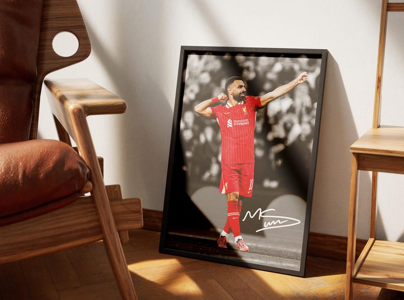 May include: Framed print of a footballer in a red kit, celebrating with arms raised. The background is a blurred black and white image. The print is in a black frame, with a signature in the bottom right corner.