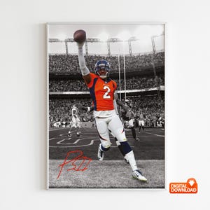 May include: A framed black and white photograph of a football player in an orange and blue uniform, holding a football above his head. The image is set against a stadium backdrop with a crowd of spectators. A red signature is at the bottom.