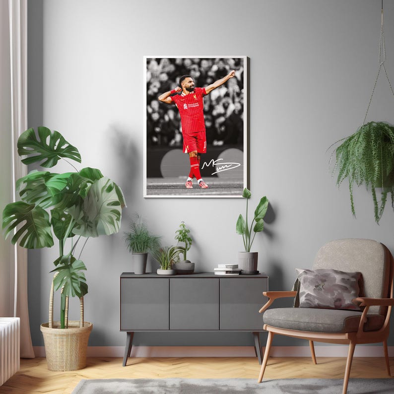 May include: A framed print of a footballer in a red kit, celebrating with arms raised. The player is on a pitch, with a crowd in the background. The print is in a white frame and has a signature.