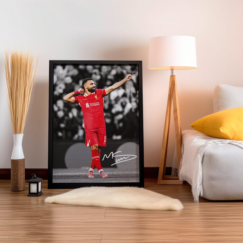 May include: Framed print of a footballer in a red kit, arms raised in celebration. The black and white background shows a crowd. The player's signature is at the bottom. The frame is black.