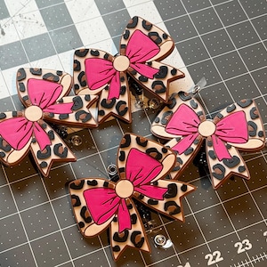 May include: Four bow-shaped badge reels with a leopard print design and bright pink bows. The badge reels have a light tan background with black and brown spots. The bows are a solid bright pink colour.