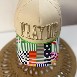 May include: A beige baseball cap with the text "PRAY BIG" embroidered in gold. The brim features a colorful patchwork design with stripes, polka dots, and squares. A green button is on top.