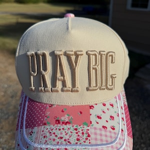 May include: A cream-colored baseball cap featuring the embroidered text PRAY BIG in a tan font. The brim displays a patchwork pattern of pink and white fabrics, including floral prints, gingham checks, and polka dots, accented by white stitching.