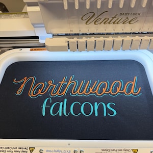 May include: A Baby Lock Venture embroidery machine with a white plastic frame holding a black fabric hoop. The fabric features the embroidered text Northwood in orange with a light blue outline, and the word falcons in light blue script.