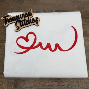 May include: White fabric with a red heart-shaped design and cursive text. The text reads "Suu" in a stylized font. A wooden cutout with the words "Treasured Stitches" is in the upper left corner.