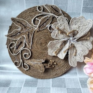 Könnte beinhalten: Ein runder, brauner Jute-Untersetzer mit floralem Design. Der Untersetzer hat eine große, cremefarbene Blume mit perlenartigen Akzenten und braunen Blättern. Der Untersetzer besteht aus gewickeltem Juteseil.