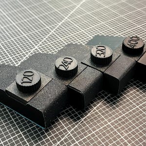 May include: Four black sanding blocks with different grit numbers: 120, 240, 320, and 400. The blocks are arranged in a row from left to right, with the highest grit number on the right.