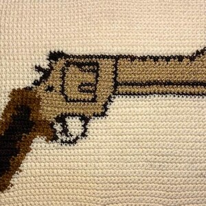 May include: A tan and brown crocheted revolver design on a cream background. The gun has a detailed barrel, cylinder, and handle, with a dark outline. The background has a textured, woven appearance.