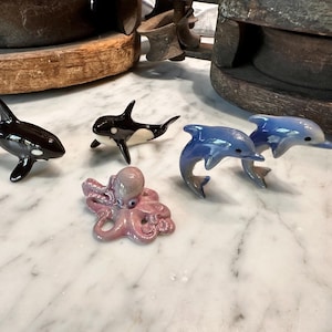 Può includere: Una collezione di figurine di animali marini in ceramica. Il set comprende due orche nere e bianche, due delfini blu e un polpo rosa. Le figurine sono esposte su una superficie di marmo bianco.