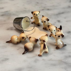 Könnte beinhalten: Eine bezaubernde Szene mit Miniatur-Keramik-Kätzchen, die einen verschütteten Eimer Sahne umgeben. Die Kätzchen haben beige und braune Markierungen mit schwarzen Akzenten. Der Eimer ist silberfarben und die Sahne hellbeige.