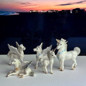 May include: A collection of white ceramic unicorn figurines with gold accents. Some have wings and blue accents. The unicorns are posed in various positions, including standing and reclining. The background shows a sunset over a beach.