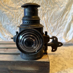 Peut inclure: Une lanterne de train vintage en métal noir avec une lentille en verre clair et un support en métal. La lanterne est de forme ronde et possède une petite cheminée sur le dessus.