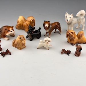 Op de afbeelding: Een verzameling keramische hondenbeeldjes in verschillende rassen en maten. De honden zijn beschilderd in realistische kleuren, waaronder bruin, zwart, wit en beige. Sommige honden staan, terwijl andere zitten of liggen. De beeldjes worden getoond op een lichtgrijze ondergrond.