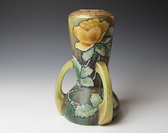 Op de afbeelding: Een gele en groene keramische vaas met twee handvatten en een bloemenontwerp. De vaas is versierd met een gele roos en groene bladeren.