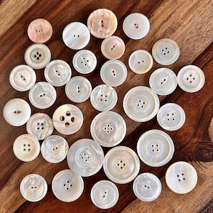 May include: A collection of various white and iridescent buttons with two or four holes, scattered on a wooden surface. The buttons vary in size and shape, with some featuring a pearlescent sheen.